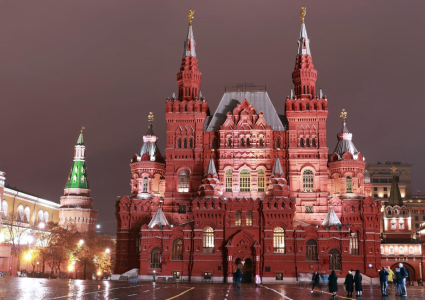 Night view of Moscow’s Red Square featuring the illuminated red-brick State Historical Museum with ornate towers and spires, wet pavement reflecting lights, and a few people walking in the foreground.