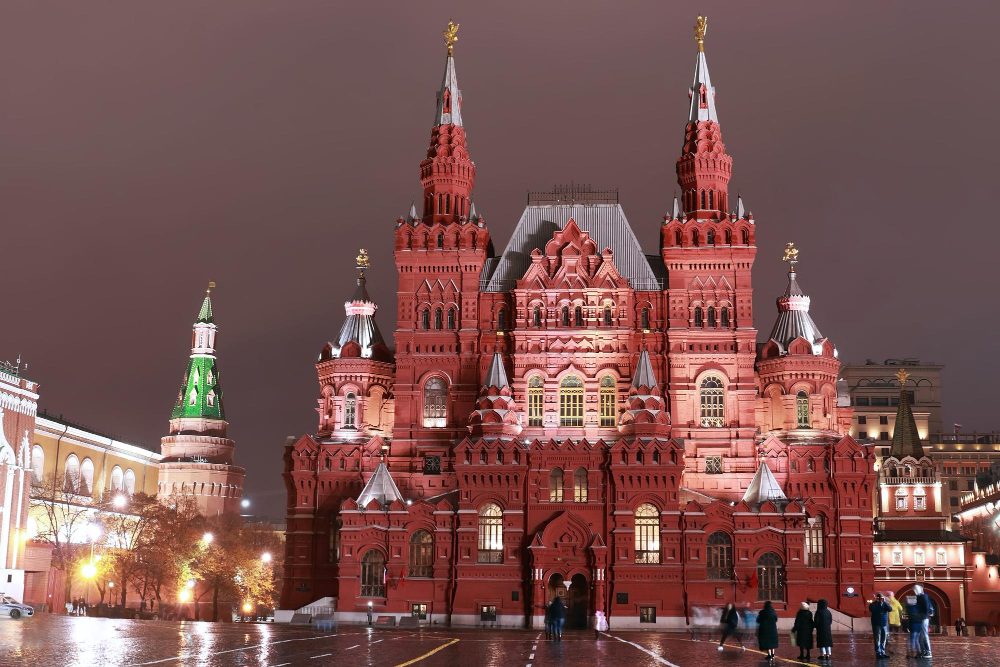 Night view of Moscow’s Red Square featuring the illuminated red-brick State Historical Museum with ornate towers and spires, wet pavement reflecting lights, and a few people walking in the foreground.